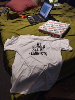 A casual setting featuring a bed with a laptop, a white T-shirt with the text 'WE SHOULD ALL BE FEMINISTS', a pizza box, and a game controller. The bed has a green cover and pillows, creating a cozy atmosphere.