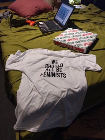 A casual setting featuring a bed with a laptop, a white T-shirt with the text 'WE SHOULD ALL BE FEMINISTS', a pizza box, and a game controller. The bed has a green cover and pillows, creating a cozy atmosphere.
