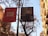 Banners hang on a pole along a street bordered by trees and buildings. One banner displays a text advertisement for a Feliu Elias exhibit at the Museu Nacional d’Art de Catalunya. The other banner shows an art piece featuring a figure leaning against a table. The trees are bare, suggesting autumn or winter, and the buildings have classic European architectural features with balconies.
