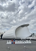 A modern architectural structure with a curved, white exterior against a cloudy sky. The foreground features a sign with the word 'Luxembourg' in both Latin and Arabic script, and the colors red, white, and blue.