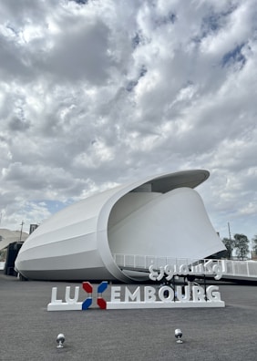 A modern architectural structure with a curved, white exterior against a cloudy sky. The foreground features a sign with the word 'Luxembourg' in both Latin and Arabic script, and the colors red, white, and blue.