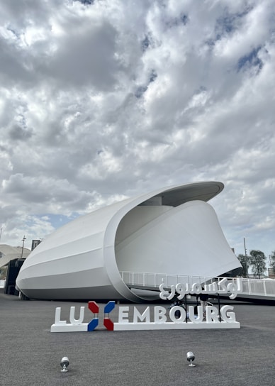 A modern architectural structure with a curved, white exterior against a cloudy sky. The foreground features a sign with the word 'Luxembourg' in both Latin and Arabic script, and the colors red, white, and blue.