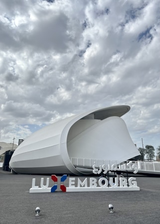 A modern architectural structure with a curved, white exterior against a cloudy sky. The foreground features a sign with the word 'Luxembourg' in both Latin and Arabic script, and the colors red, white, and blue.