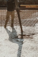 A construction worker is pouring wet concrete over a grid of rebar on a construction site. The worker is wearing boots and is holding a large hose that dispenses the concrete onto the surface. Shadows are cast on the textured surface as the concrete spreads out.
