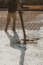 A construction worker is pouring wet concrete over a grid of rebar on a construction site. The worker is wearing boots and is holding a large hose that dispenses the concrete onto the surface. Shadows are cast on the textured surface as the concrete spreads out.