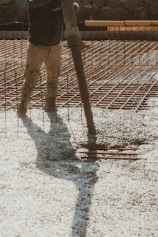 A construction worker is pouring wet concrete over a grid of rebar on a construction site. The worker is wearing boots and is holding a large hose that dispenses the concrete onto the surface. Shadows are cast on the textured surface as the concrete spreads out.
