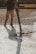 A construction worker is pouring wet concrete over a grid of rebar on a construction site. The worker is wearing boots and is holding a large hose that dispenses the concrete onto the surface. Shadows are cast on the textured surface as the concrete spreads out.