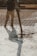 A construction worker is pouring wet concrete over a grid of rebar on a construction site. The worker is wearing boots and is holding a large hose that dispenses the concrete onto the surface. Shadows are cast on the textured surface as the concrete spreads out.