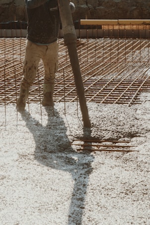 A construction worker is pouring wet concrete over a grid of rebar on a construction site. The worker is wearing boots and is holding a large hose that dispenses the concrete onto the surface. Shadows are cast on the textured surface as the concrete spreads out.