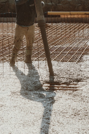 A construction worker is pouring wet concrete over a grid of rebar on a construction site. The worker is wearing boots and is holding a large hose that dispenses the concrete onto the surface. Shadows are cast on the textured surface as the concrete spreads out.