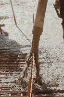Concrete is being poured onto a construction site through a large hose. Rebar is visible, indicating reinforcement work for a foundation or slab. The surrounding area appears organized, with construction tools partially visible.