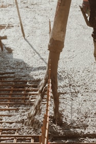 Concrete is being poured onto a construction site through a large hose. Rebar is visible, indicating reinforcement work for a foundation or slab. The surrounding area appears organized, with construction tools partially visible.