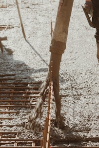 Concrete is being poured onto a construction site through a large hose. Rebar is visible, indicating reinforcement work for a foundation or slab. The surrounding area appears organized, with construction tools partially visible.