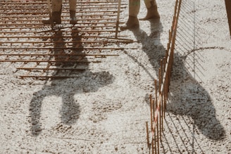 A shadow of a person standing over a grid of metal reinforcement bars on a construction site. The ground is covered with gravel, and the lighting creates strong shadows.