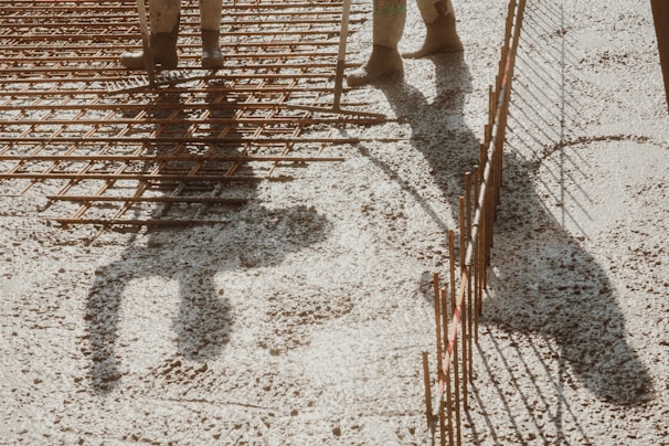 A shadow of a person standing over a grid of metal reinforcement bars on a construction site. The ground is covered with gravel, and the lighting creates strong shadows.