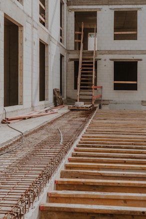 A partially constructed building featuring concrete walls and openings for windows. A staircase leads to an unfinished upper level, with construction materials and wooden beams scattered around.