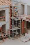 A construction site featuring scaffolding erected against a partially built structure. The building is made of light-colored concrete blocks and red bricks. The scaffolding is metal and multi-tiered, providing access to different levels. Construction materials such as wooden planks and metal bars are visible. The area is dusty and appears to be in the early stages of construction.