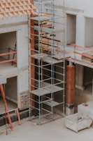 A construction site featuring scaffolding erected against a partially built structure. The building is made of light-colored concrete blocks and red bricks. The scaffolding is metal and multi-tiered, providing access to different levels. Construction materials such as wooden planks and metal bars are visible. The area is dusty and appears to be in the early stages of construction.