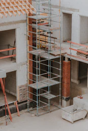 A construction site featuring scaffolding erected against a partially built structure. The building is made of light-colored concrete blocks and red bricks. The scaffolding is metal and multi-tiered, providing access to different levels. Construction materials such as wooden planks and metal bars are visible. The area is dusty and appears to be in the early stages of construction.