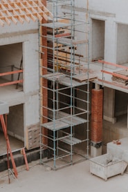 A construction site featuring scaffolding erected against a partially built structure. The building is made of light-colored concrete blocks and red bricks. The scaffolding is metal and multi-tiered, providing access to different levels. Construction materials such as wooden planks and metal bars are visible. The area is dusty and appears to be in the early stages of construction.