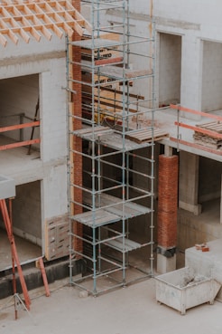 A construction site featuring scaffolding erected against a partially built structure. The building is made of light-colored concrete blocks and red bricks. The scaffolding is metal and multi-tiered, providing access to different levels. Construction materials such as wooden planks and metal bars are visible. The area is dusty and appears to be in the early stages of construction.