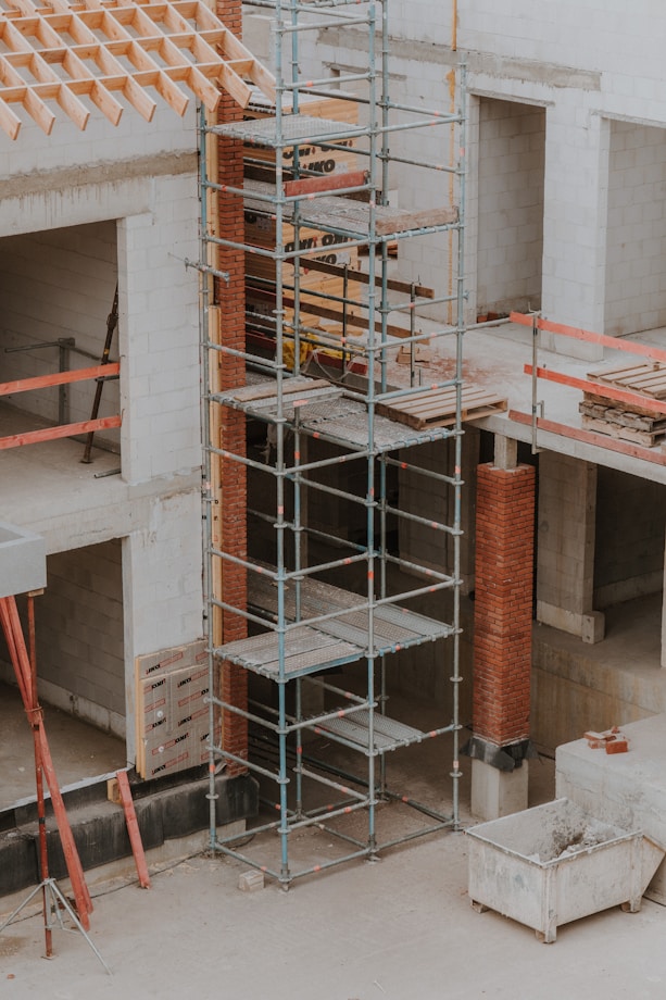 A construction site featuring scaffolding erected against a partially built structure. The building is made of light-colored concrete blocks and red bricks. The scaffolding is metal and multi-tiered, providing access to different levels. Construction materials such as wooden planks and metal bars are visible. The area is dusty and appears to be in the early stages of construction.