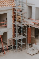 A construction site featuring scaffolding erected against a partially built structure. The building is made of light-colored concrete blocks and red bricks. The scaffolding is metal and multi-tiered, providing access to different levels. Construction materials such as wooden planks and metal bars are visible. The area is dusty and appears to be in the early stages of construction.