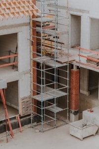 A construction site featuring scaffolding erected against a partially built structure. The building is made of light-colored concrete blocks and red bricks. The scaffolding is metal and multi-tiered, providing access to different levels. Construction materials such as wooden planks and metal bars are visible. The area is dusty and appears to be in the early stages of construction.