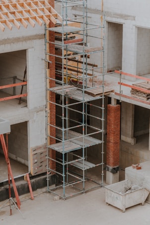 A construction site featuring scaffolding erected against a partially built structure. The building is made of light-colored concrete blocks and red bricks. The scaffolding is metal and multi-tiered, providing access to different levels. Construction materials such as wooden planks and metal bars are visible. The area is dusty and appears to be in the early stages of construction.