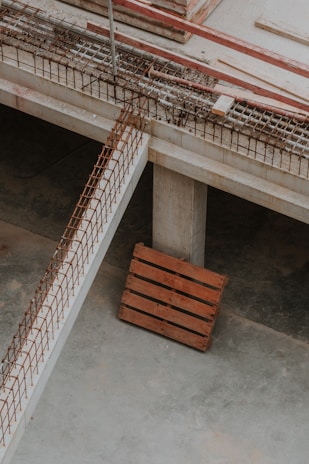 An industrial setting featuring part of a construction site. The image includes concrete structures, reinforcing steel bars arranged in a grid pattern, and a wooden pallet resting on the concrete floor. The environment has a raw, unfinished appearance indicative of an in-progress building project.