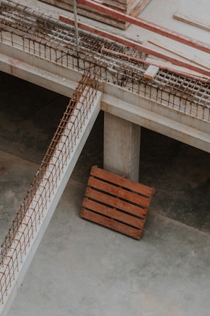 An industrial setting featuring part of a construction site. The image includes concrete structures, reinforcing steel bars arranged in a grid pattern, and a wooden pallet resting on the concrete floor. The environment has a raw, unfinished appearance indicative of an in-progress building project.