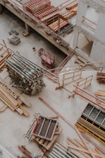 An overhead view of a construction site showing various materials and equipment organized on the ground. Wooden planks, metal poles, and scaffolding are visible along with a red motorized platform lift. The setting appears to be within a partially constructed building, with concrete floors and unfinished walls.