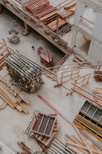 An overhead view of a construction site showing various materials and equipment organized on the ground. Wooden planks, metal poles, and scaffolding are visible along with a red motorized platform lift. The setting appears to be within a partially constructed building, with concrete floors and unfinished walls.