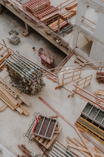 An overhead view of a construction site showing various materials and equipment organized on the ground. Wooden planks, metal poles, and scaffolding are visible along with a red motorized platform lift. The setting appears to be within a partially constructed building, with concrete floors and unfinished walls.