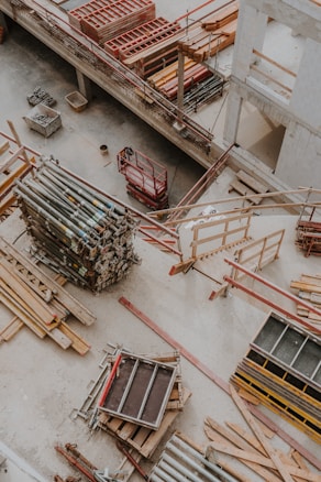 An overhead view of a construction site showing various materials and equipment organized on the ground. Wooden planks, metal poles, and scaffolding are visible along with a red motorized platform lift. The setting appears to be within a partially constructed building, with concrete floors and unfinished walls.
