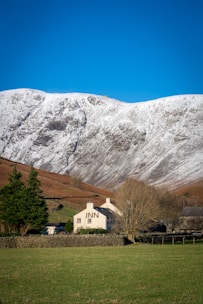 A picturesque landscape featuring a quaint inn with a traditional chimney set against the backdrop of a snow-capped mountain. The inn is surrounded by green fields and neatly arranged stone fences, with a few trees and bushes adding to the natural beauty. The sky is clear and bright blue, enhancing the serene atmosphere.