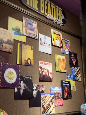 A wall display features various album covers and posters related to The Beatles and their individual members. The arrangement is colorful and artistic, showcasing different portraits and designs. A prominent sign at the top reads 'THE BEATLES Discography,' highlighting the theme of the collection.