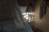 A cozy bedroom setting with soft lighting, featuring a bed with white linens and a sheer canopy. Natural light casts plant-like shadows on the wall. A woven pendant lamp hangs from the ceiling, and a small wooden stool holds an open book. Towels or fabrics are draped on wooden hooks attached to the wall.