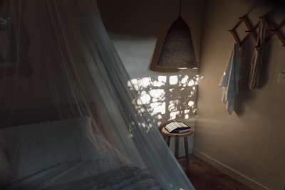 A cozy bedroom setting with soft lighting, featuring a bed with white linens and a sheer canopy. Natural light casts plant-like shadows on the wall. A woven pendant lamp hangs from the ceiling, and a small wooden stool holds an open book. Towels or fabrics are draped on wooden hooks attached to the wall.