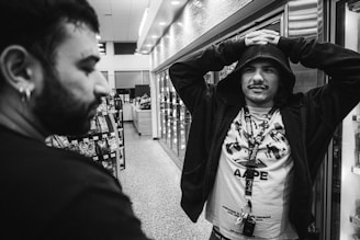 A black and white photo of two men inside a store. One man on the right is wearing a hoodie with the hood up and a shirt with a pattern of shapes and words. He has his arms raised, hands resting on his head, appearing nonchalant. The man on the left is in profile, with short hair and an earring, looking towards the other man. The background includes shelves with products and a lit ceiling, suggesting a retail environment.