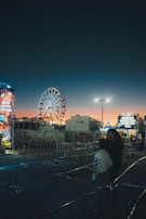 A brightly illuminated Ferris wheel stands in the middle of a fairground at dusk, with colorful lights and signs advertising various food stalls, including bacon themed ones. The sky transitions from a deep twilight blue to a warm orange near the horizon. Two people are seen in the foreground, standing by a barricade.