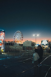 A brightly illuminated Ferris wheel stands in the middle of a fairground at dusk, with colorful lights and signs advertising various food stalls, including bacon themed ones. The sky transitions from a deep twilight blue to a warm orange near the horizon. Two people are seen in the foreground, standing by a barricade.