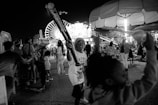 A lively nighttime scene at an amusement park featuring children playing with inflatable bats. A large Ferris wheel is illuminated in the background, and various rides and attractions are visible. Many people are mingling and enjoying the festive atmosphere.
