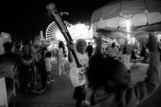 A lively nighttime scene at an amusement park featuring children playing with inflatable bats. A large Ferris wheel is illuminated in the background, and various rides and attractions are visible. Many people are mingling and enjoying the festive atmosphere.