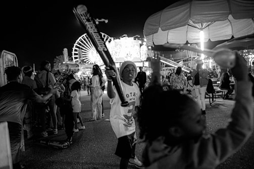 A lively nighttime scene at an amusement park featuring children playing with inflatable bats. A large Ferris wheel is illuminated in the background, and various rides and attractions are visible. Many people are mingling and enjoying the festive atmosphere.