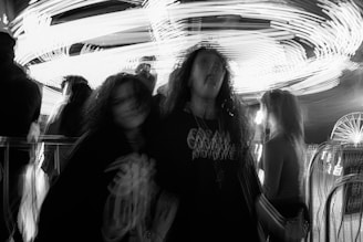 A black and white image of a carnival scene featuring blurred figures, possibly in motion, set against the backdrop of bright lights and a ferris wheel. The motion blur suggests energy and movement.