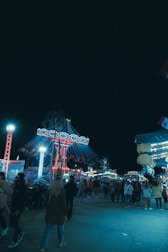 A lively amusement park scene at night. Brightly lit rides, including a carousel with swinging seats, are surrounded by a crowd of people enjoying the festive atmosphere. The scene is bustling with activity and colorful lights illuminate the area.