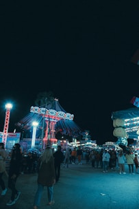 A lively amusement park scene at night. Brightly lit rides, including a carousel with swinging seats, are surrounded by a crowd of people enjoying the festive atmosphere. The scene is bustling with activity and colorful lights illuminate the area.