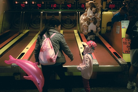 A woman and a child are at a skee-ball arcade game. The woman is bending down, possibly explaining the game or helping the child. The child is holding a pink dolphin balloon and is wearing a pink hoodie and pink cap. Behind them, there are plush toys hanging as prizes and the arcade machine displays various scores.