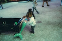 A child holding a large green and pink mermaid doll, standing next to a black cart with toys inside. Other people can be seen walking in the background on a textured concrete surface.